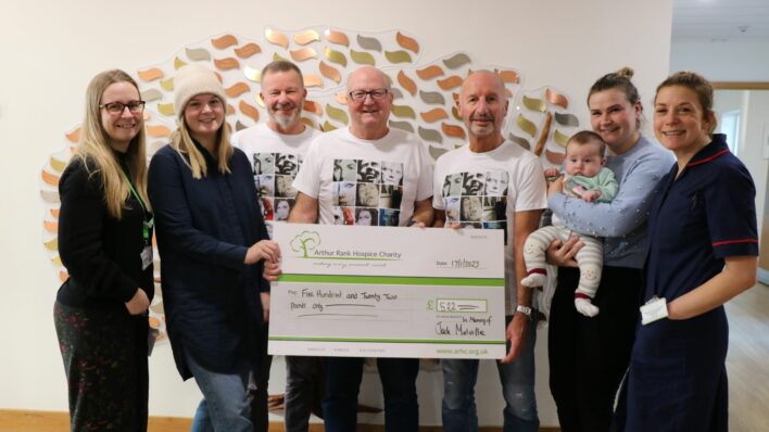 Cheque being held by 3 men with 4 women one holding a baby one in nurse uniform in front of memory tree