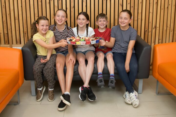 4 girls and 1 boy holding a tray of cakes sitting on a sofa
