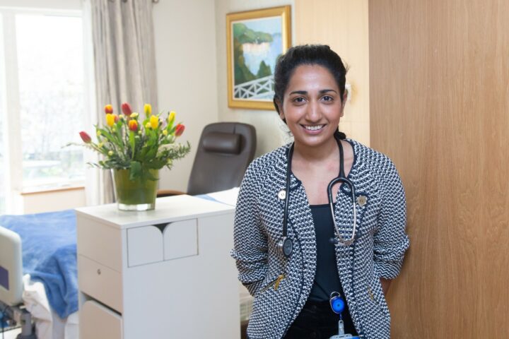 Female Dr with stethoscope smiling in front of bed and flowers