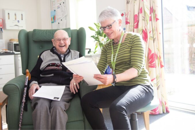Male in an armchair smiling and female holding a sheet of white paper