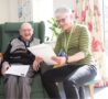 Male in an armchair smiling and female holding a sheet of white paper