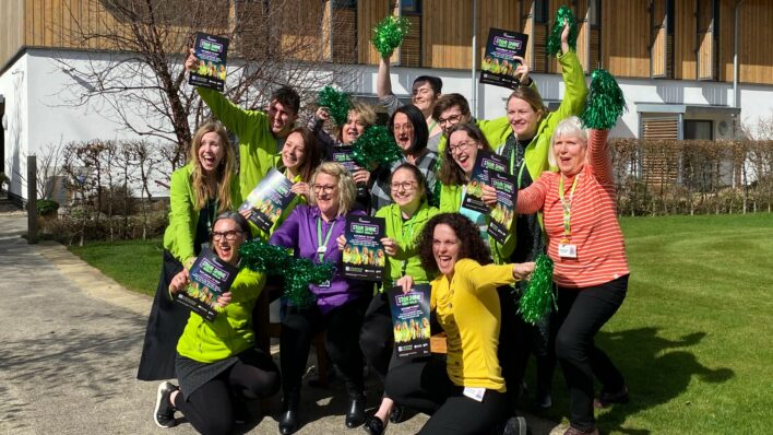 Group 1 male and females wearing mainly green holding flyers and green pom poms smiling at the camera