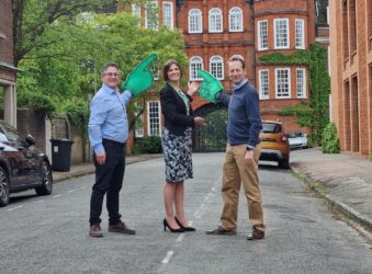 2 males and 1 female standing in the street holding large green foam fingers