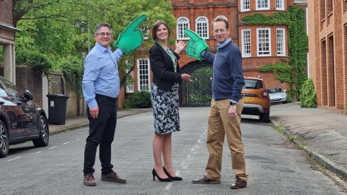 2 males and 1 female standing in the street holding large green foam fingers