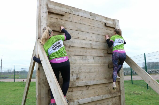 two females wearing branded t-shirts climbing a wooden wall