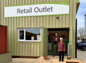 Man and woman standing at the door step of a shop, smiling at camera. The sign of the shop reads "Retail Outlet".