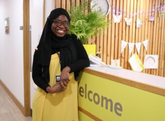 Female wearing black headress and yellow dress smiling at the camera leaning on reception desk