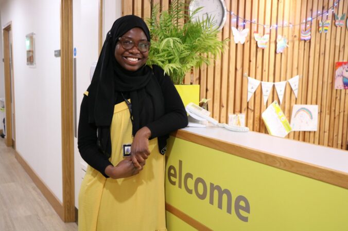 Female wearing black headress and yellow dress smiling at the camera leaning on reception desk