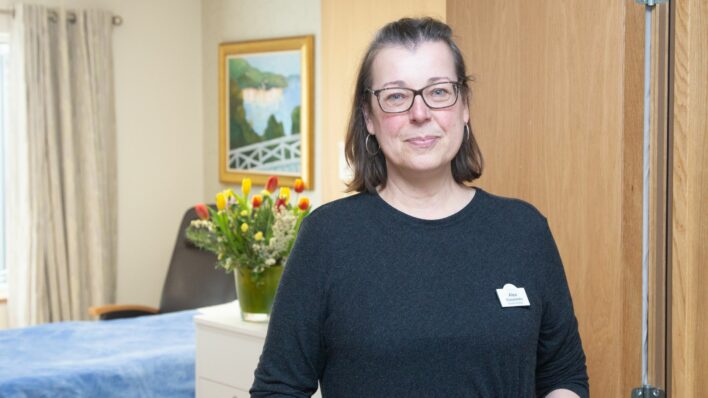 Female wearing glasses standing in front of a bed a vase of flowers