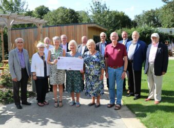 Group of men and women holding a large cheque in the garden in front of the shed