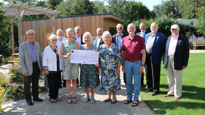 Group of men and women holding a large cheque in the garden in front of the shed