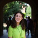 Female wearing green t-shirt standing in front of an arch outside, smiling at the camera 