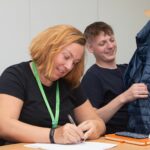 Female sitting at a desk writing and a male next to her holding a coat up