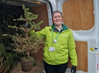 Lady in a green jacket standing next to a van and holding a small, shabby Christmas tree