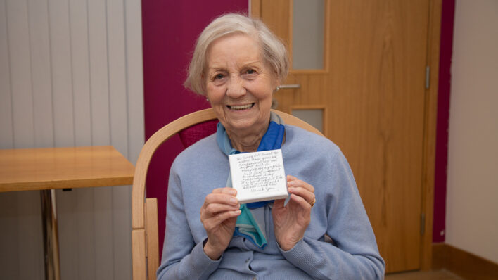 Female sitting on a chair holding a small canvas, smiling at the camera