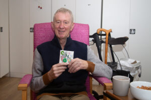 Male sitting in a pink chair, holding a small canvas, looking at the camera. 