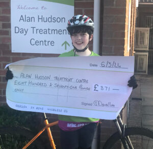 Young boy on a bike with green branded top holding a large cheque