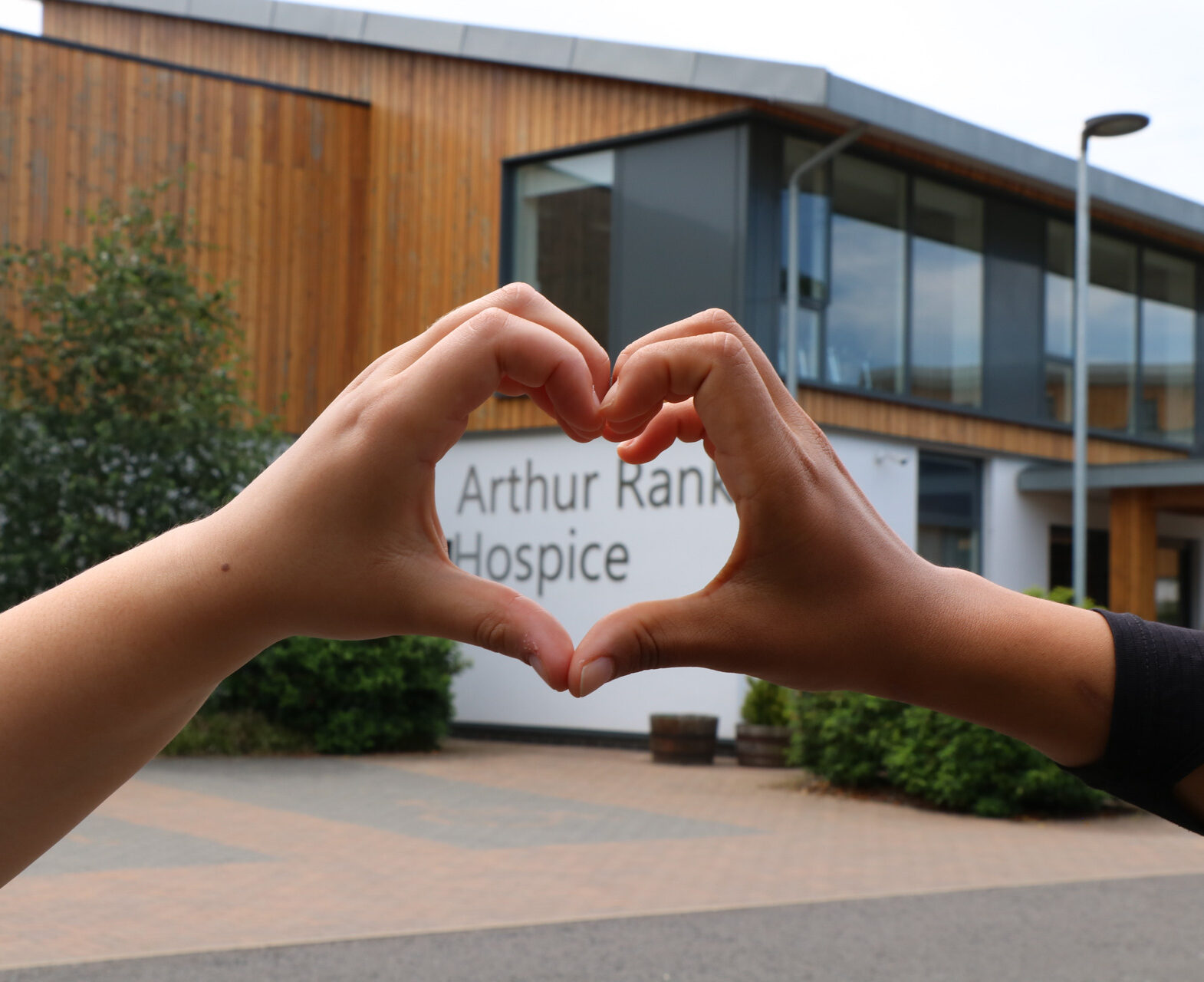 2 hands making a heart shape in front of building and sign Arthur Rank Hospice