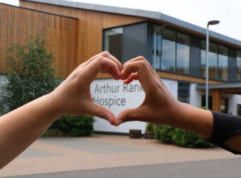 2 hands making a heart shape in front of building and sign Arthur Rank Hospice