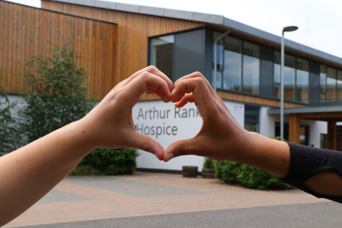 2 hands making a heart shape in front of building and sign Arthur Rank Hospice