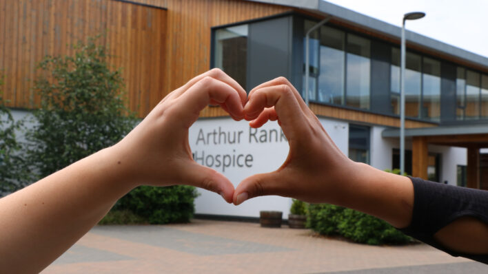 2 hands making a heart shape in front of building and sign Arthur Rank Hospice