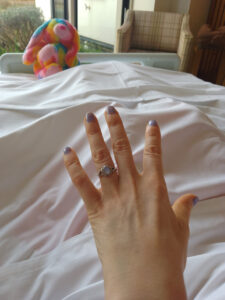 A hand wearing a ring and rainbow teddy at the end of a bed