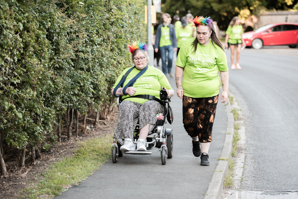 Lady on the route in an electric wheelchair wearing green tshirt and multicoloured wig on hair. Alongside her is a lady walking, wearing green tshirt and multicoloured wig in hair. 
