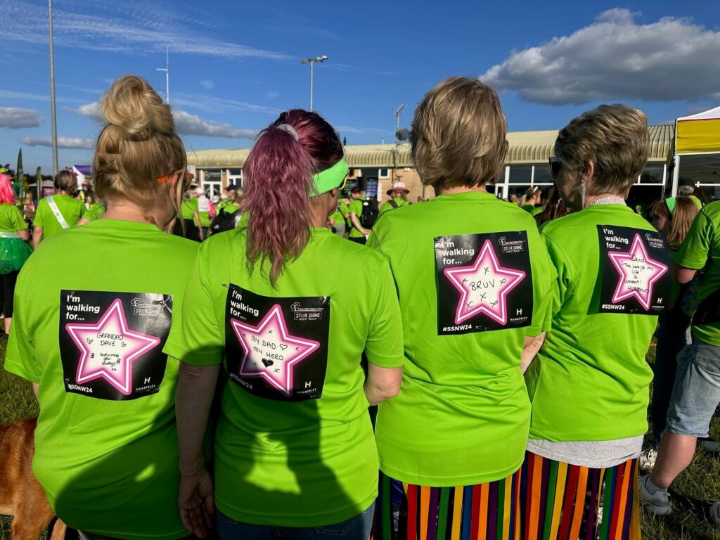 Four ladies standing with their backs to the camera. They are wearing green tshirts and have in memory labels on their backs.