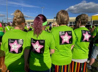 Four ladies standing with their backs to the camera. They are wearing green tshirts and have in memory labels on their backs.