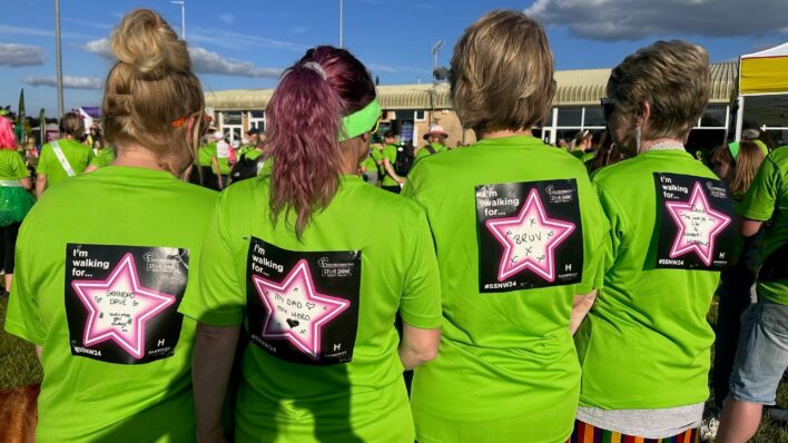 Four ladies standing with their backs to the camera. They are wearing green tshirts and have in memory labels on their backs.