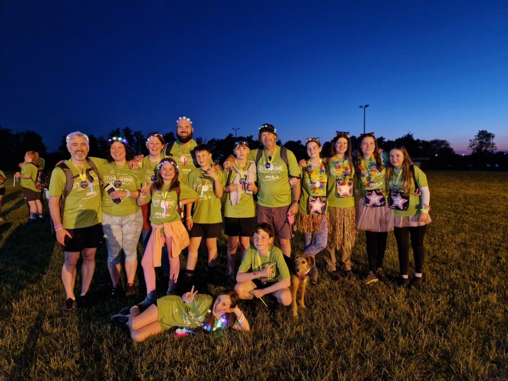 Group of people wearing green tshirts, huddled tigether in celebratory photo. The darkness of the sky behind them illuminates the image of them.