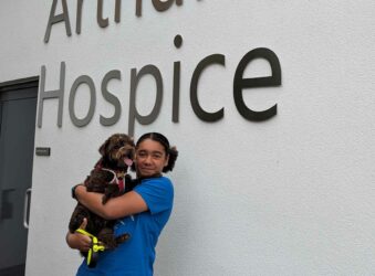image on young girl in blue top stood behind a white wall with hospice written on, holding brown dog