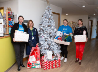 4 females (including one nurse) standing in front of a silver Christmas Tree, holding gifts.