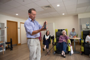 Male standing with an object and 3 females in chairs looking 