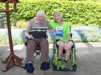 Outside in the sun older man and a younger lady hugging, both in wheelchairs