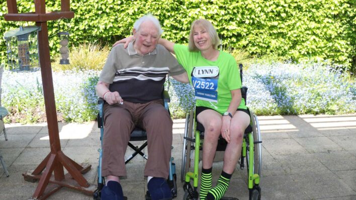Outside in the sun older man and a younger lady hugging, both in wheelchairs