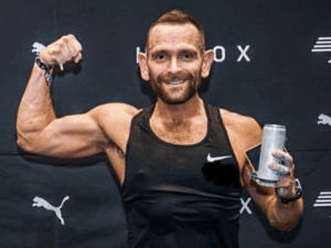 man flexing arm muscle smiling at camera on Hyrox background