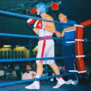 Man boxing in ring wearing white next to trainer in blue shirt