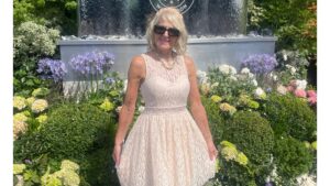 Blonde lady in sunglasses cream dress in front of Wimbledon sign and flowers