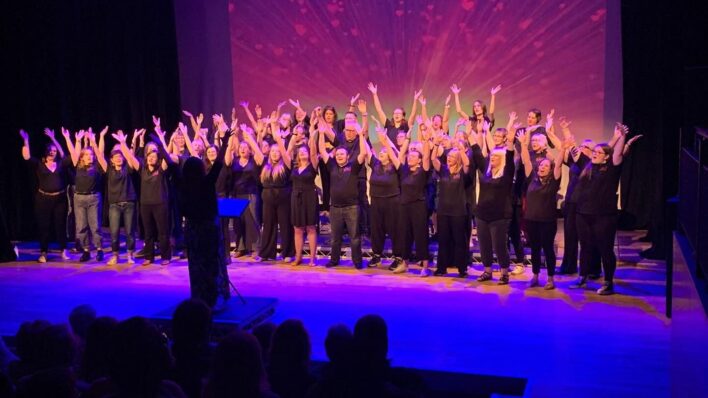 40+ people on a stage dressed in Black, singing Stage light up blue and purple.
