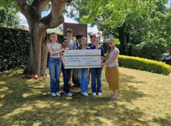 Outside under a tree 7 people looking at camera with giant cheque