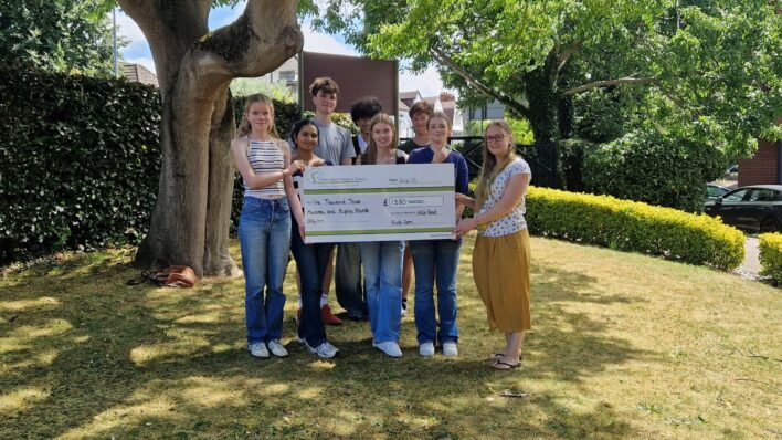 Outside under a tree 7 people looking at camera with giant cheque