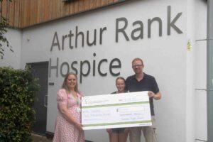 2 females and male holding a large cheque, standing outside Arthur Rank Hospice lettering