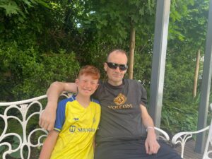 boy in yellow sitting next to man in black t-shirt on outdoor bench at Arthur Rank