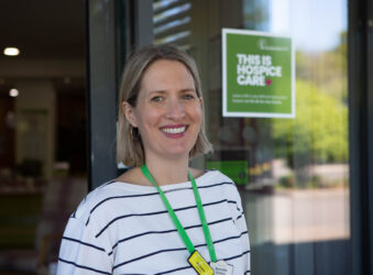 A blond lady in front of a glass door, wearing white top with black stripes and a green lanyard.