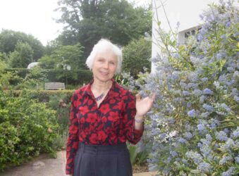 Person standing in a garden beside flowering shrubs, wearing a red patterned top and raising one hand.