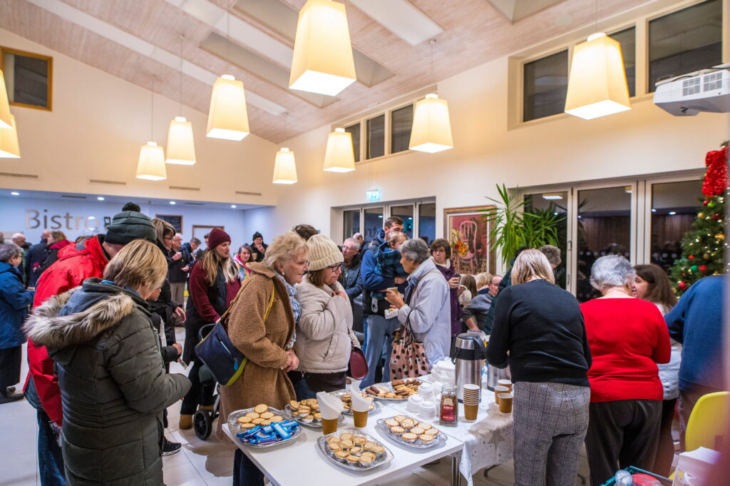 Several people mingling around a table set out with hot drinks and mince pies.